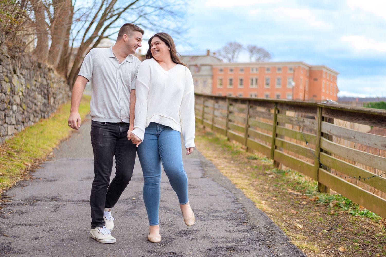 Couple walking together on path beside rustic wooden fence and brick building.