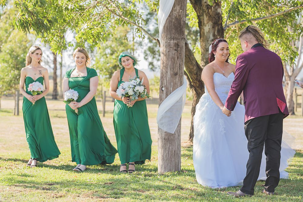 Wedding party in green dresses and formal attire poses for photos outdoors under trees with beautiful lighting.