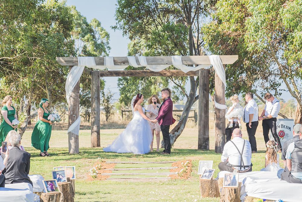 People gather outdoors under large trees for a wedding ceremony with white chairs and floral decorations.