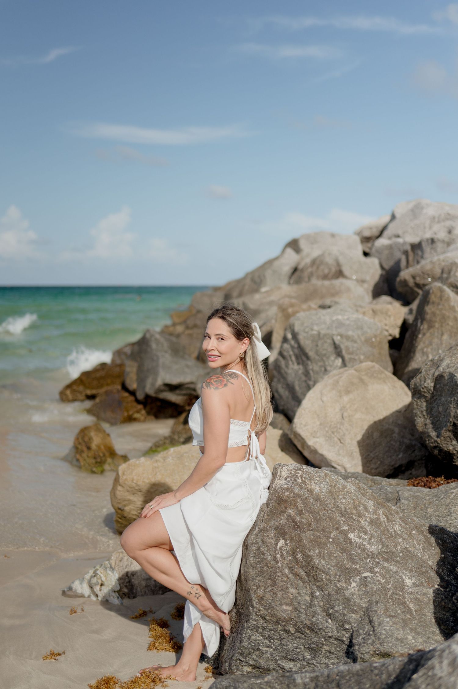 A person in a white dress poses on coastal rocks with turquoise ocean waves and blue sky in the background.