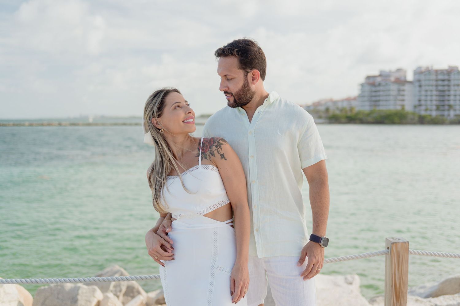 Couple in white outfits embraces and smiles at each other on a pier with turquoise ocean and buildings in background.
