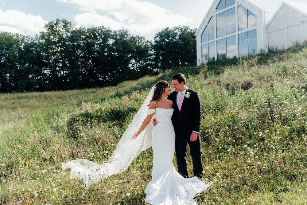 A couple in wedding attire embraces on a grassy hillside with a modern white barn venue in the background.
