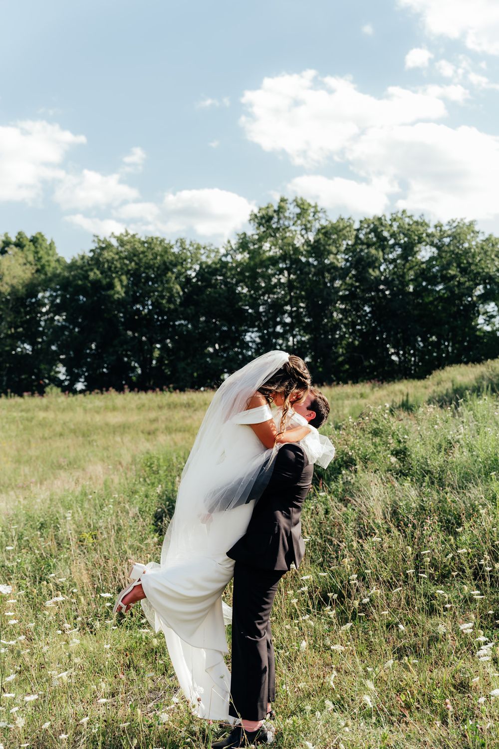 Newlywed couple embraces in a sunlit meadow with flowing white wedding dress and trees in background.