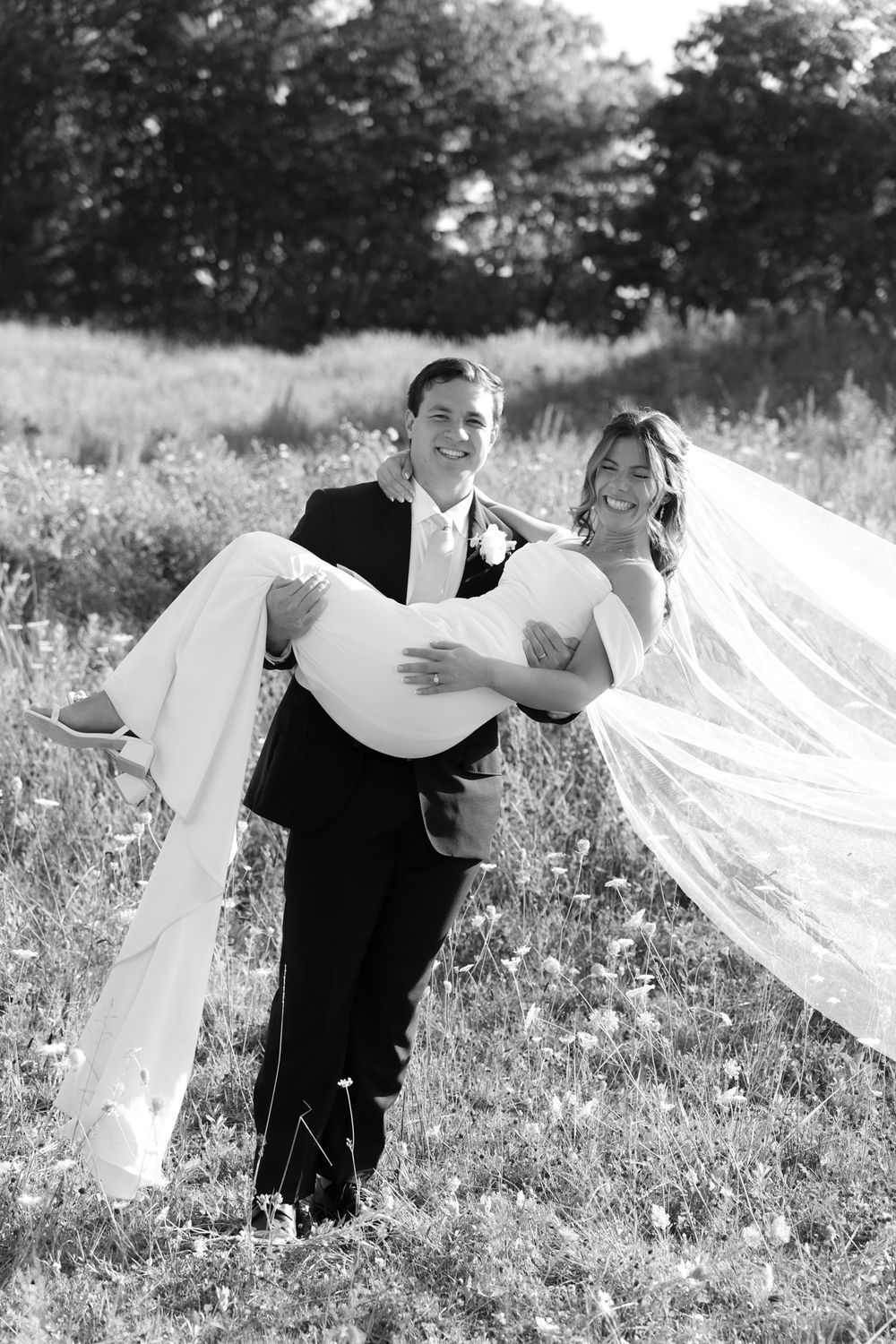 A black and white wedding photo of a couple in a field with the groom carrying the bride as her veil flows.