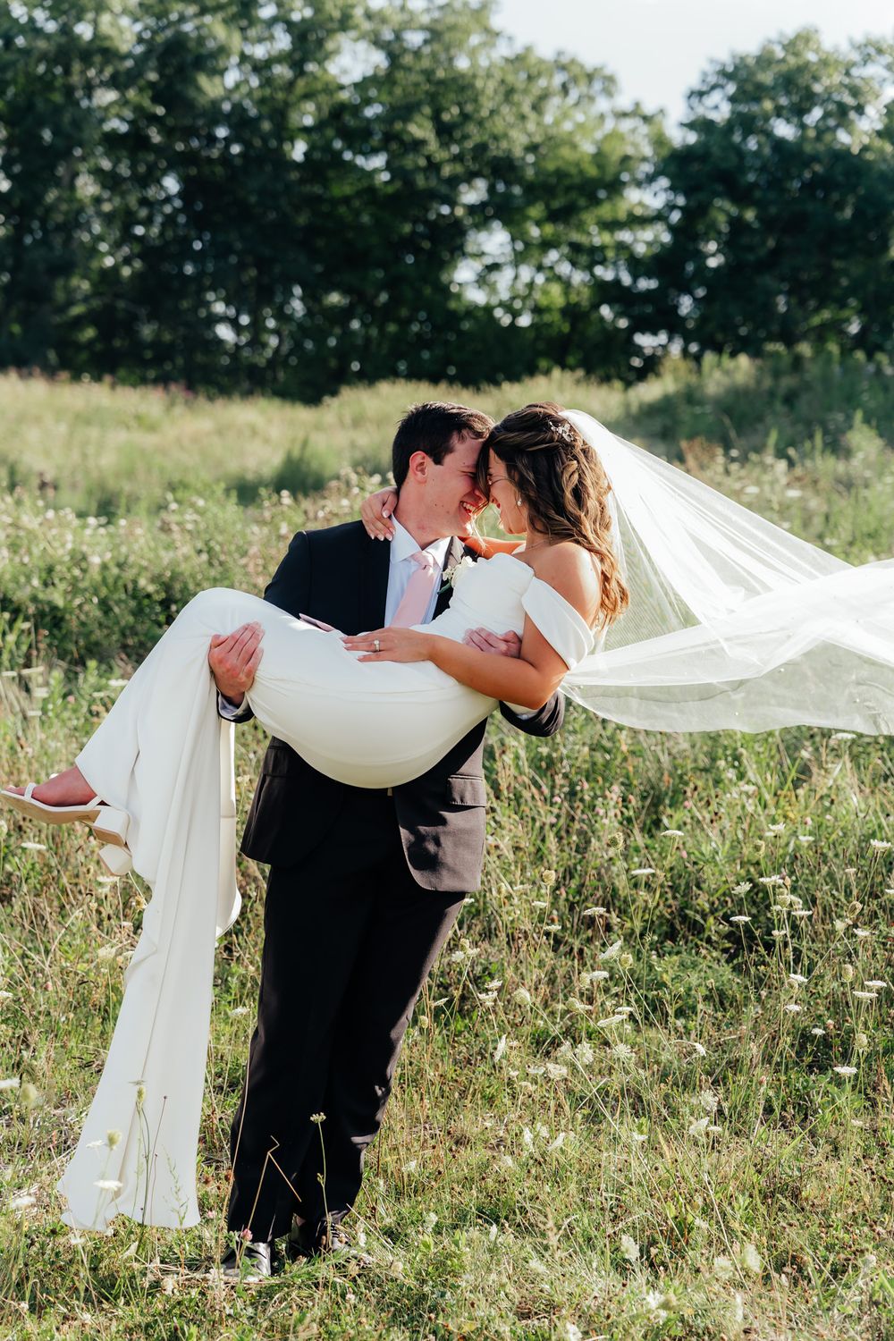 A romantic wedding portrait of a groom carrying his bride in a flowing white dress across a sunlit meadow.