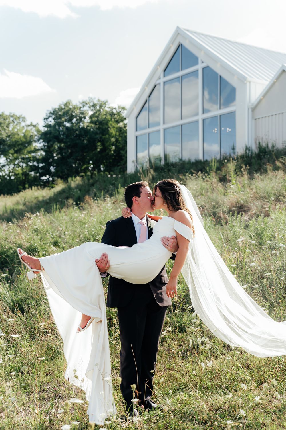 A romantic wedding couple embrace in a wildflower field with a modern white chapel in the background.