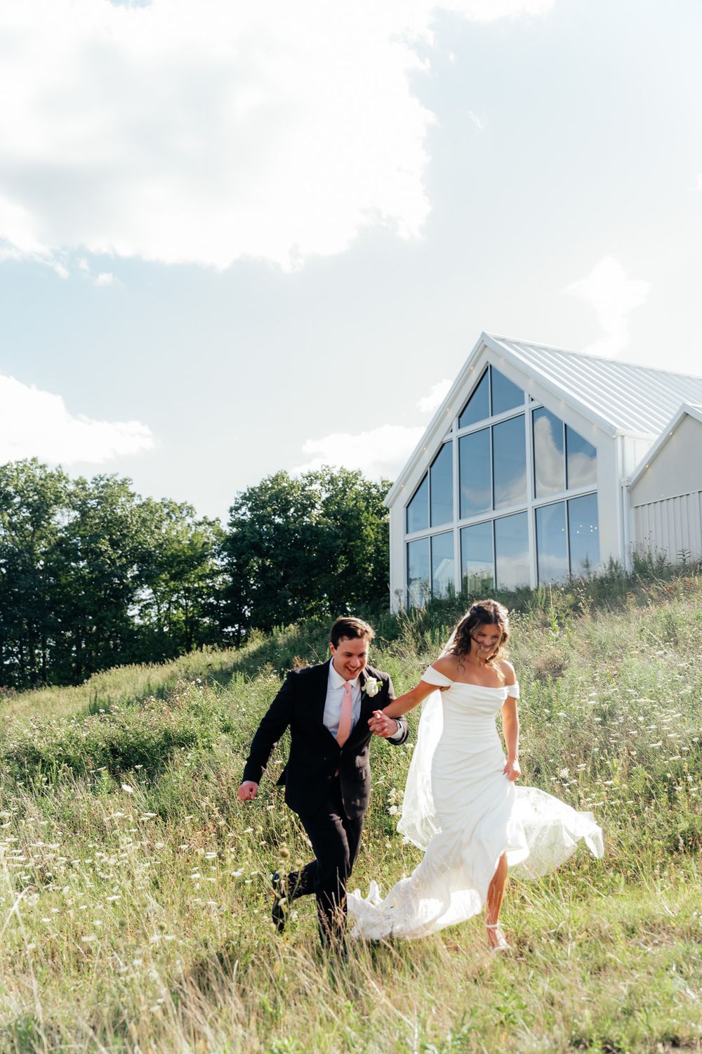 A couple runs joyfully through a grassy hillside with a modern white chapel building in the background.