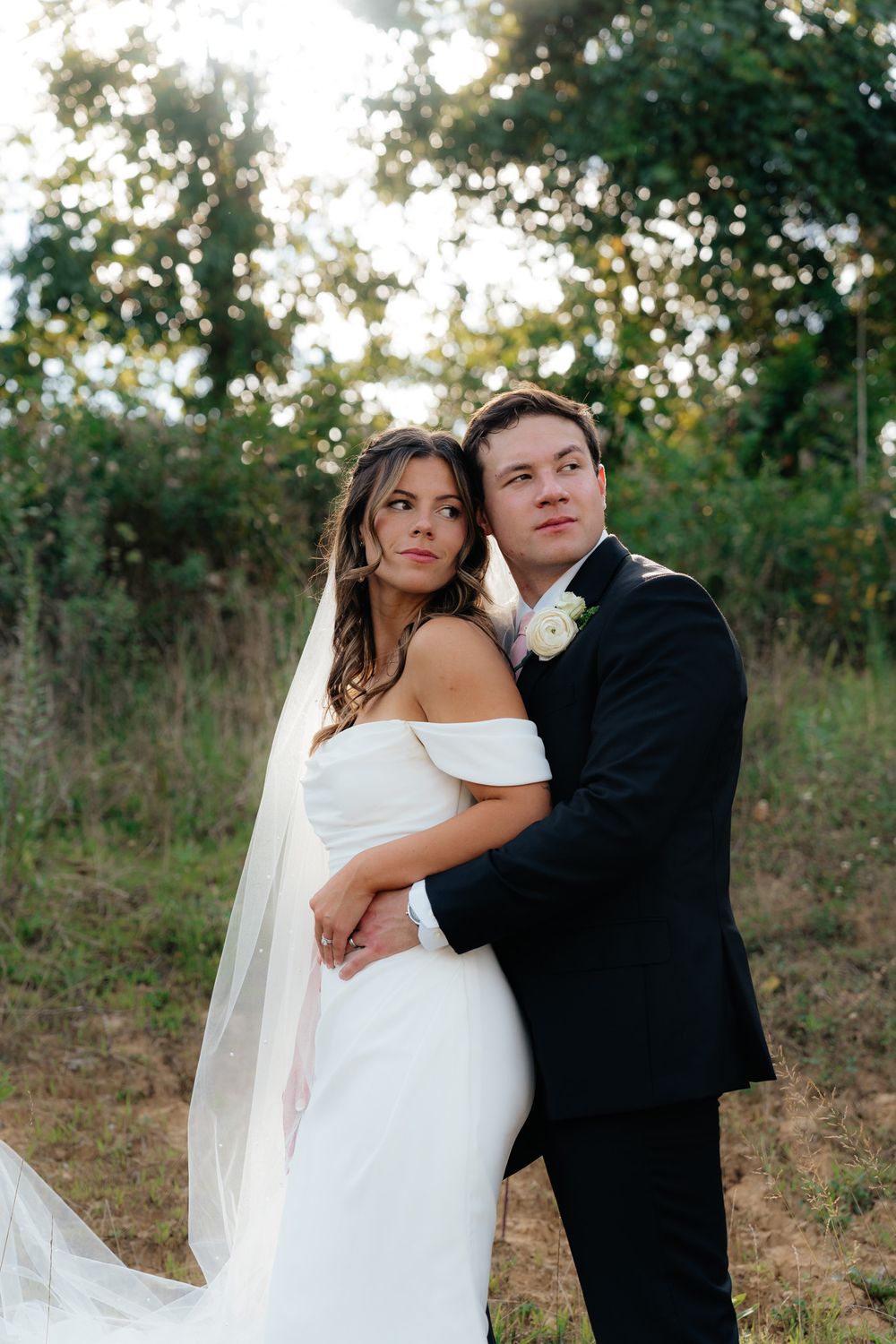A couple poses together outdoors for their wedding photos, wearing classic white gown and black tuxedo in a natural setting.