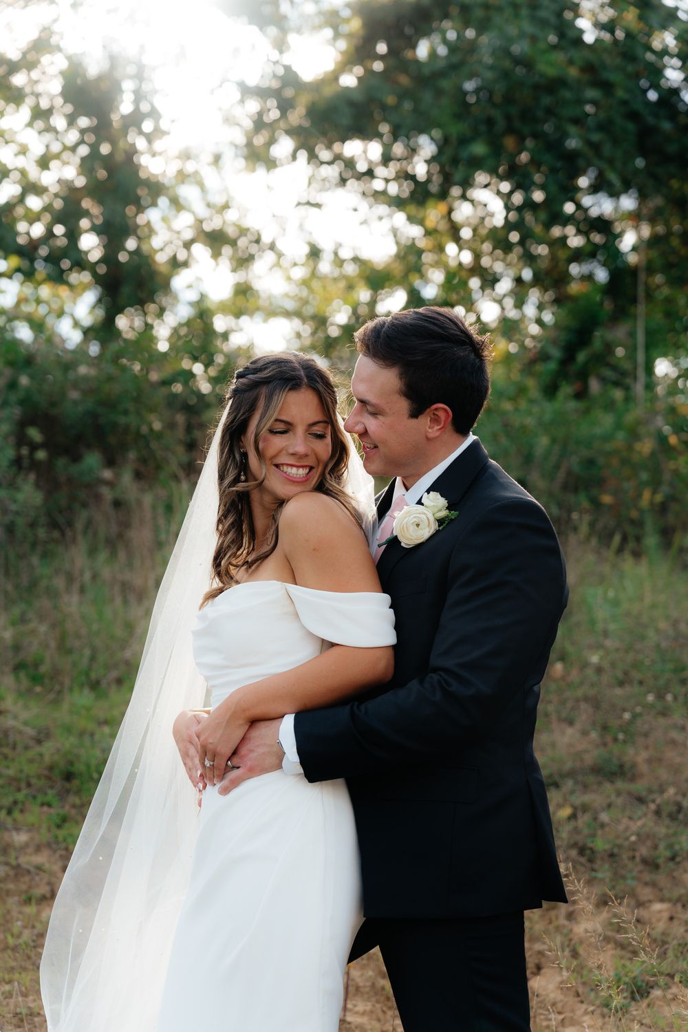 Romantic wedding couple embraces outdoors during golden hour with sunlit trees in background.