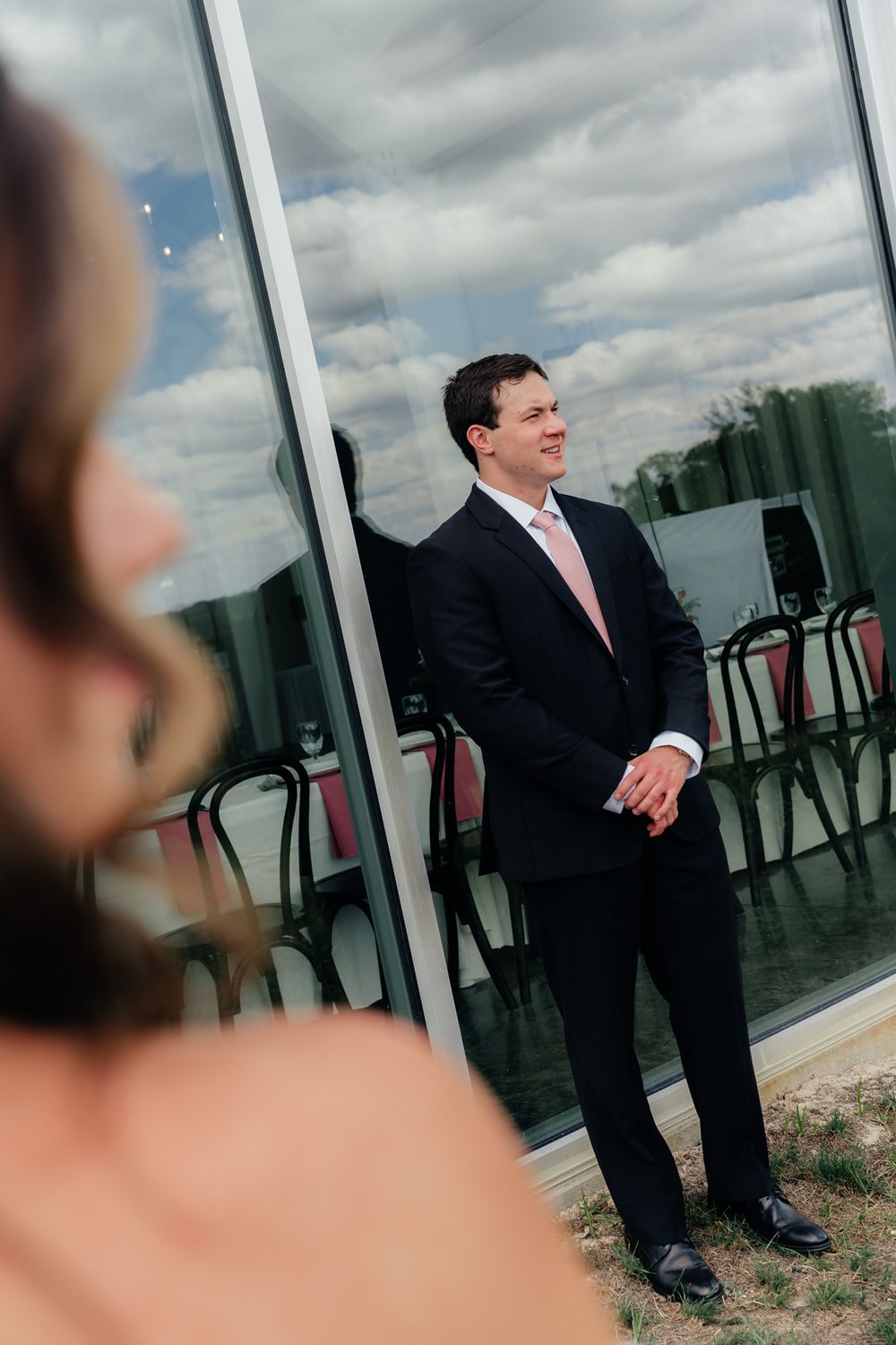 A professionally dressed person in a dark suit and pink tie stands outdoors against a cloudy sky with chairs visible nearby.