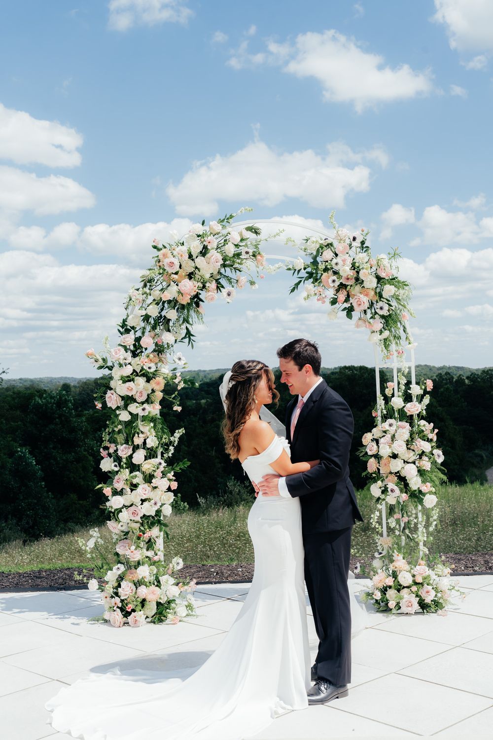 A romantic wedding ceremony under a beautiful floral arch with pink and white roses against a blue summer sky.