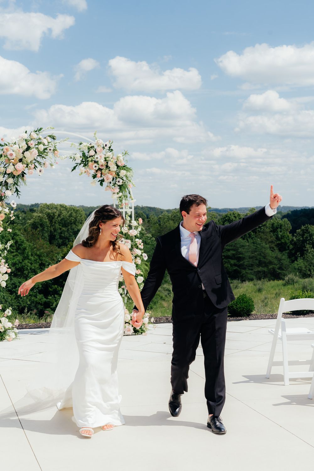 A joyful wedding couple dances on an outdoor terrace with a floral arch and scenic mountain views in the background.