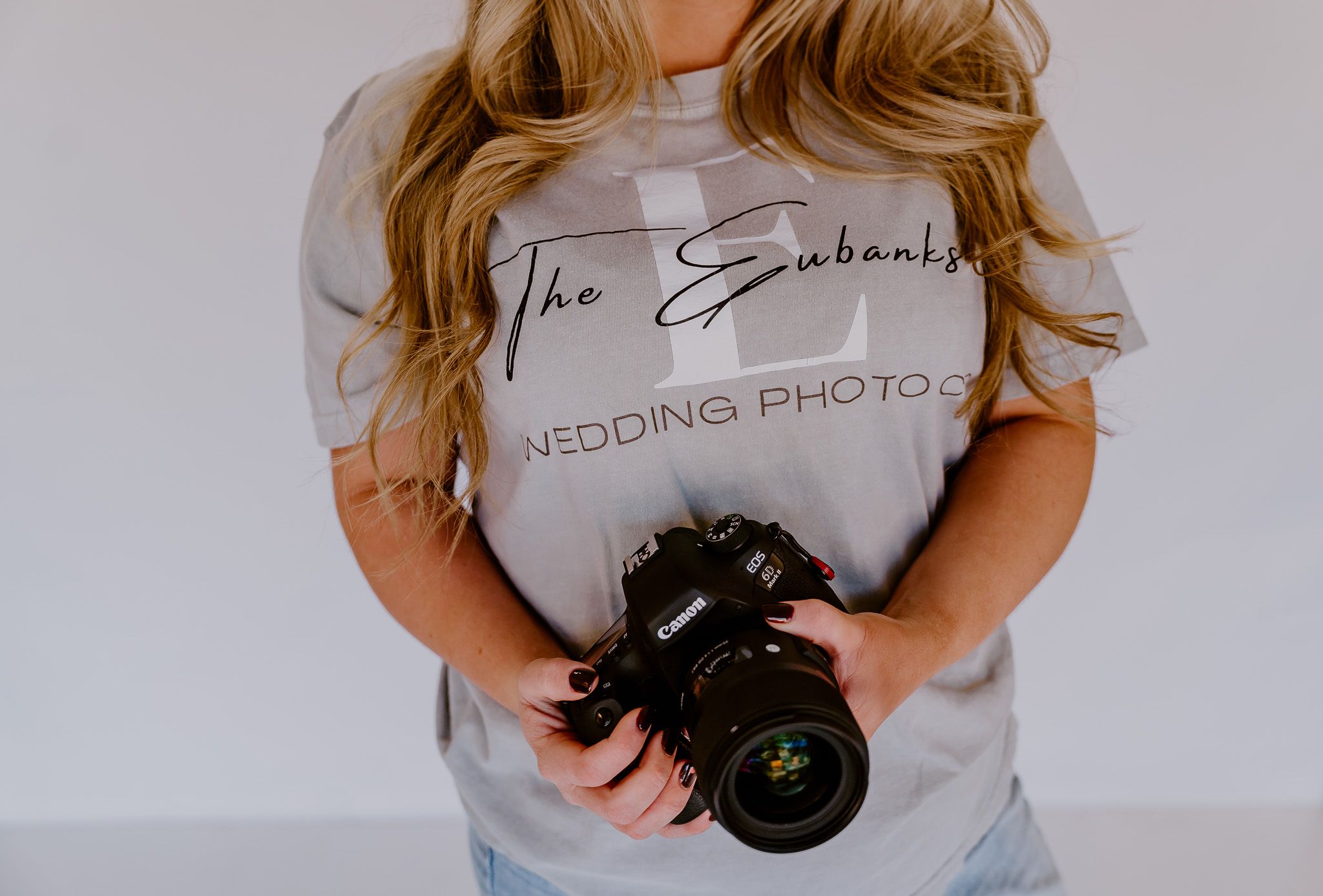 Photographer wearing a light grey "The Eyesmith Wedding Photo" t-shirt holds a professional DSLR camera.