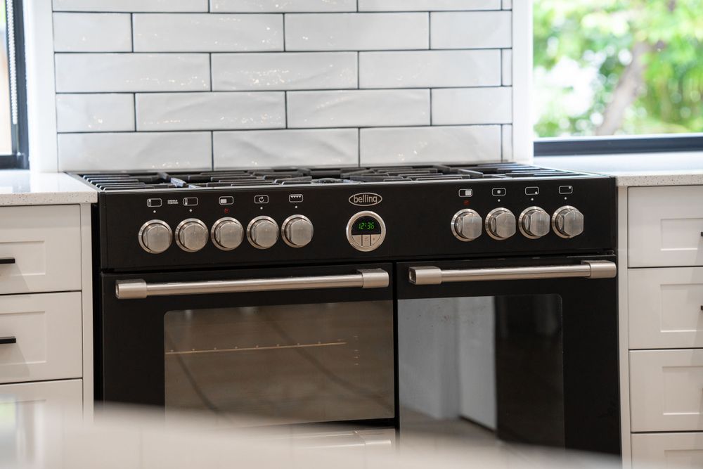 A close-up shot of a sleek, black, stainless steel oven with gas burners, set against a white tiled backsplash and bright kitchen surroundin