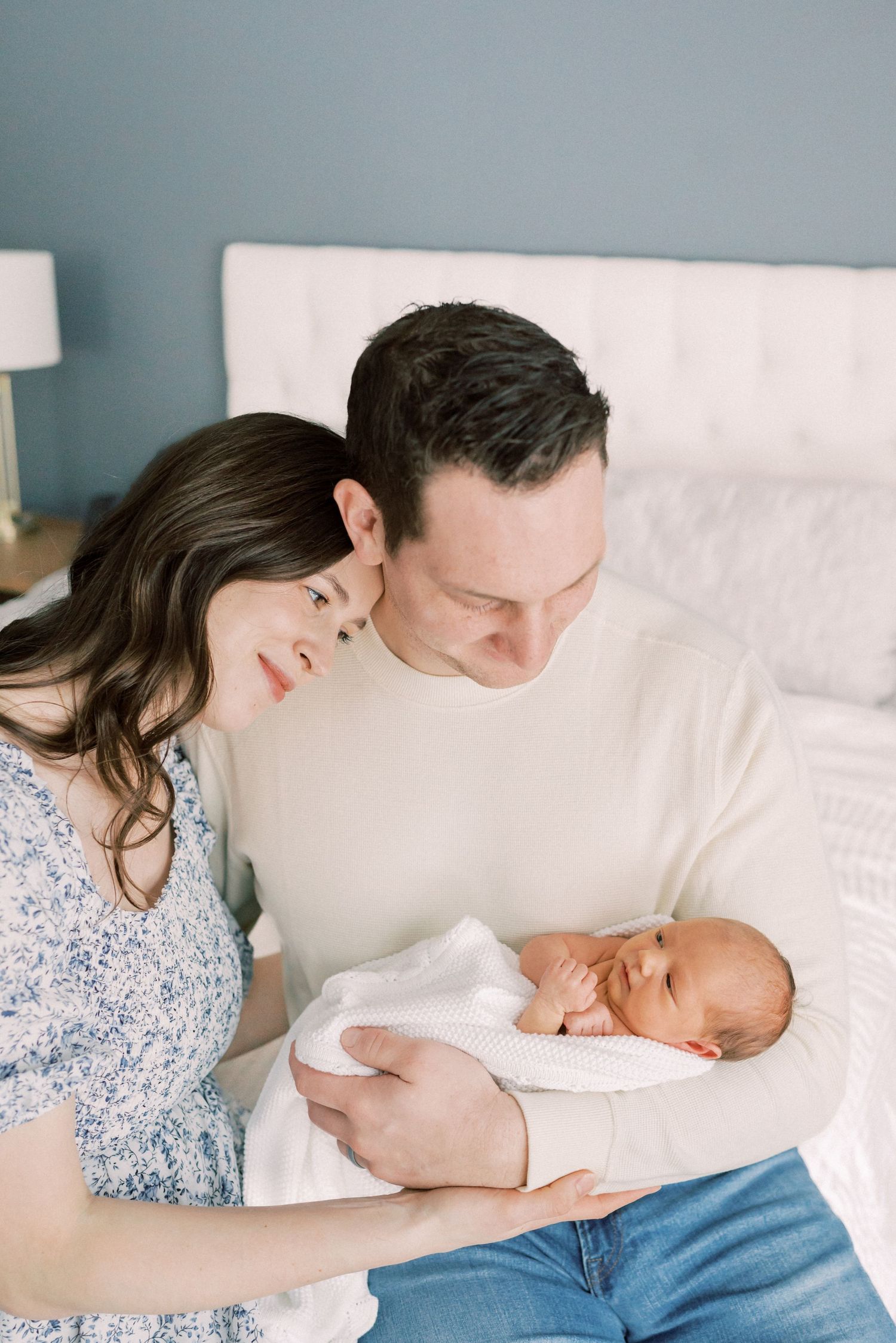Parents share tender moment with newborn baby while resting together on white bedding.