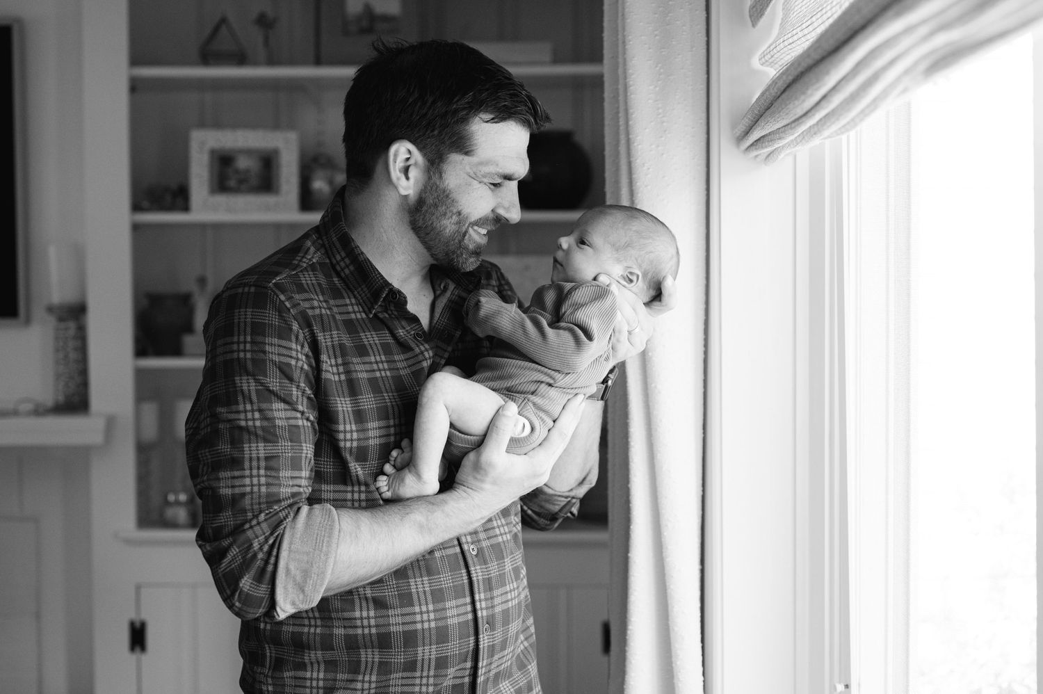 A tender black and white moment between parent and newborn baby looking out a sunlit window.