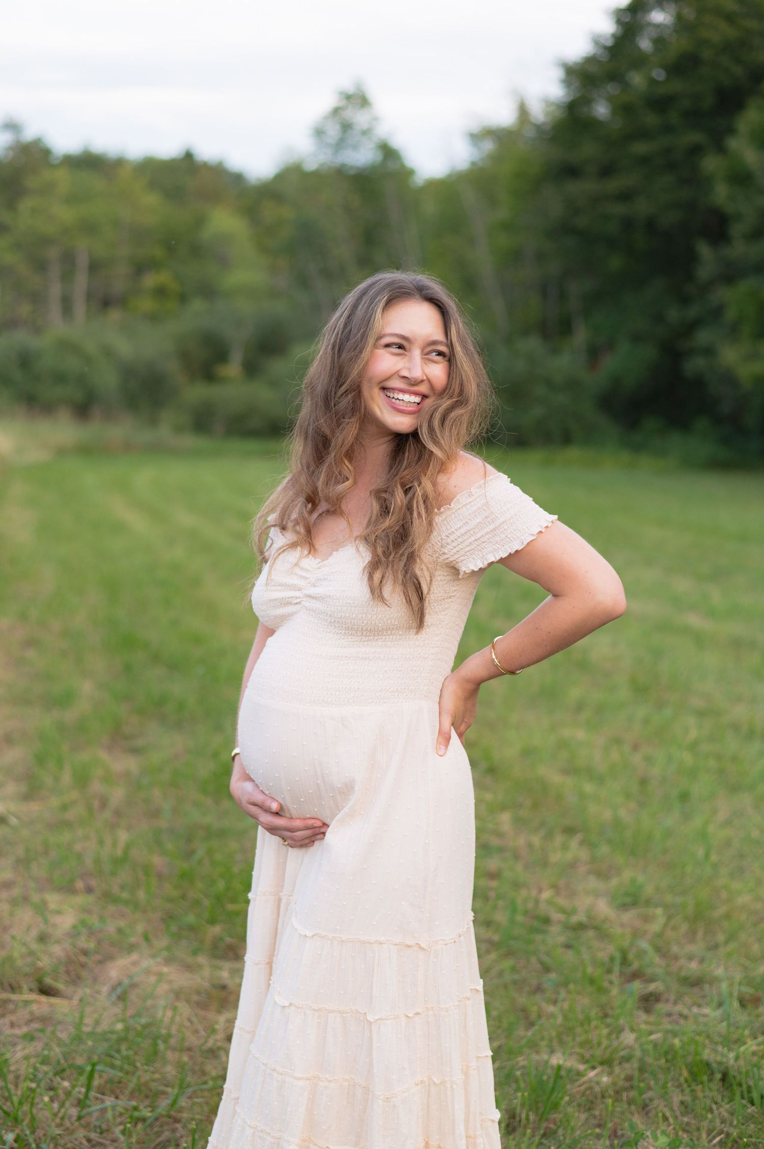 A pregnant woman in a flowy off-white dress poses outdoors in a grassy field with trees in the background.