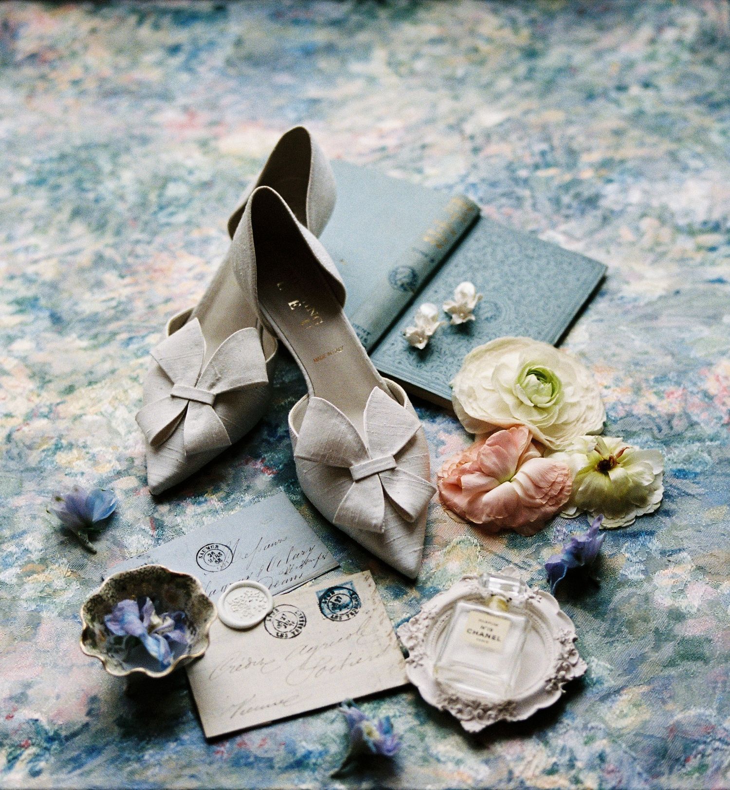Vintage wedding accessories including beige bow heels, delicate roses, and elegant invitation details on a textured surface.