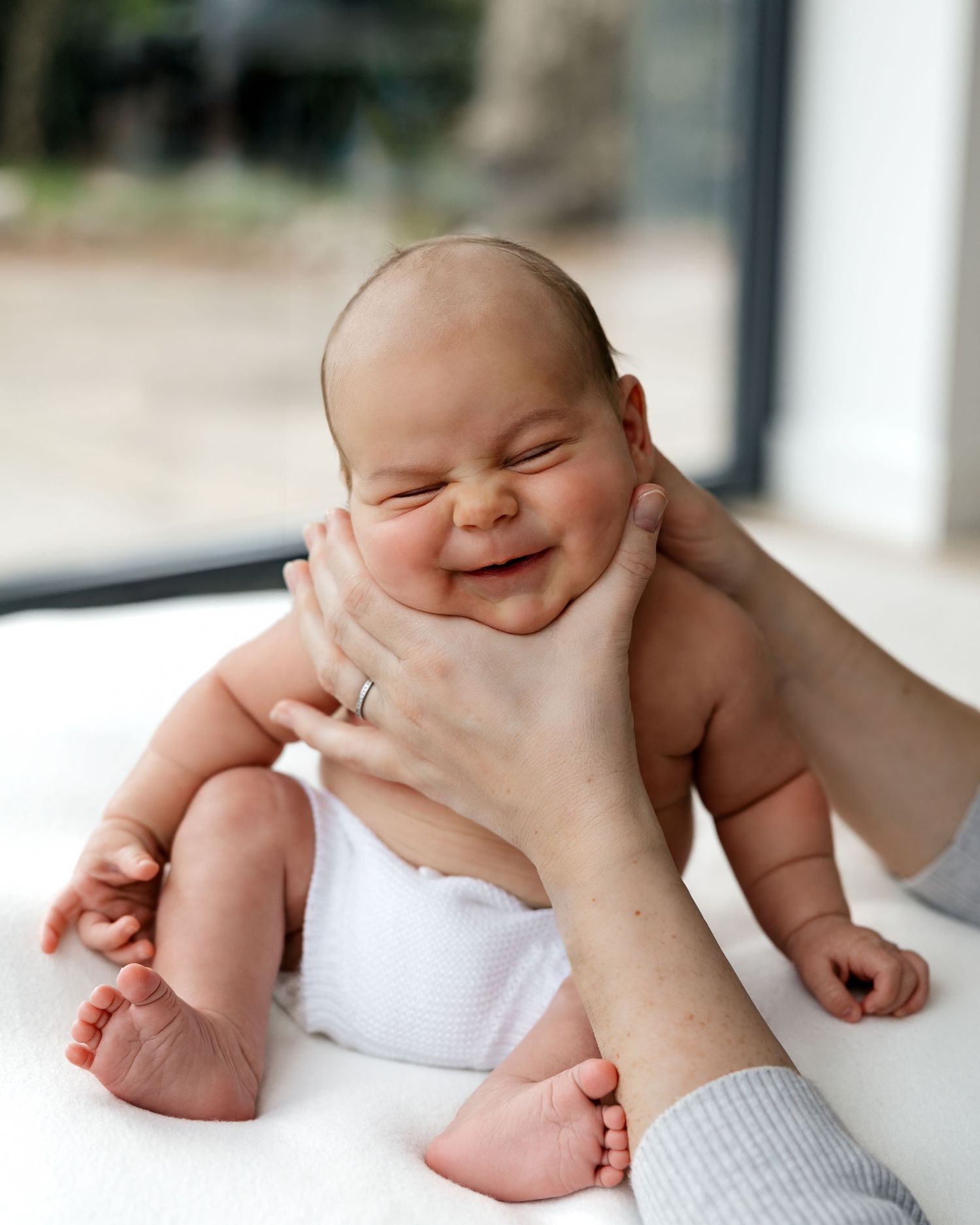 Adorable baby in a white cloth smiles with pure joy while being held by gentle hands.