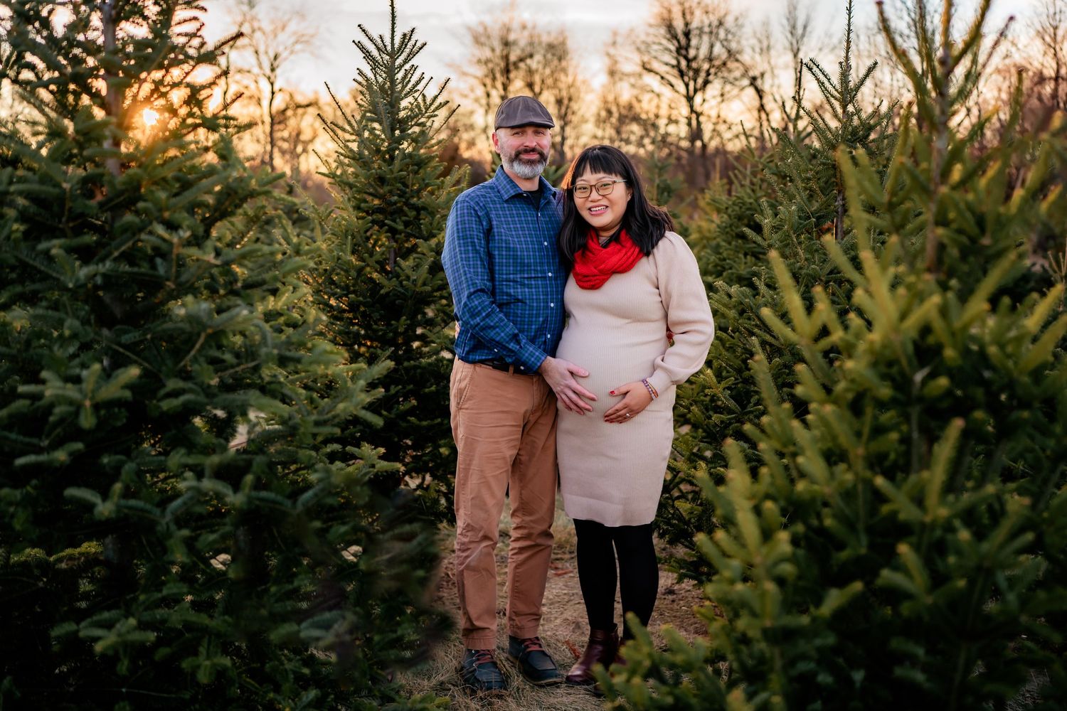 A couple stands together at a Christmas tree farm during sunset, surrounded by evergreen trees.