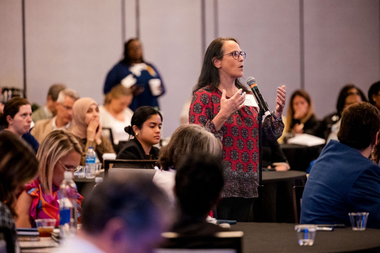 A person stands to speak at a professional conference with other attendees seated at round tables in a large meeting room.
