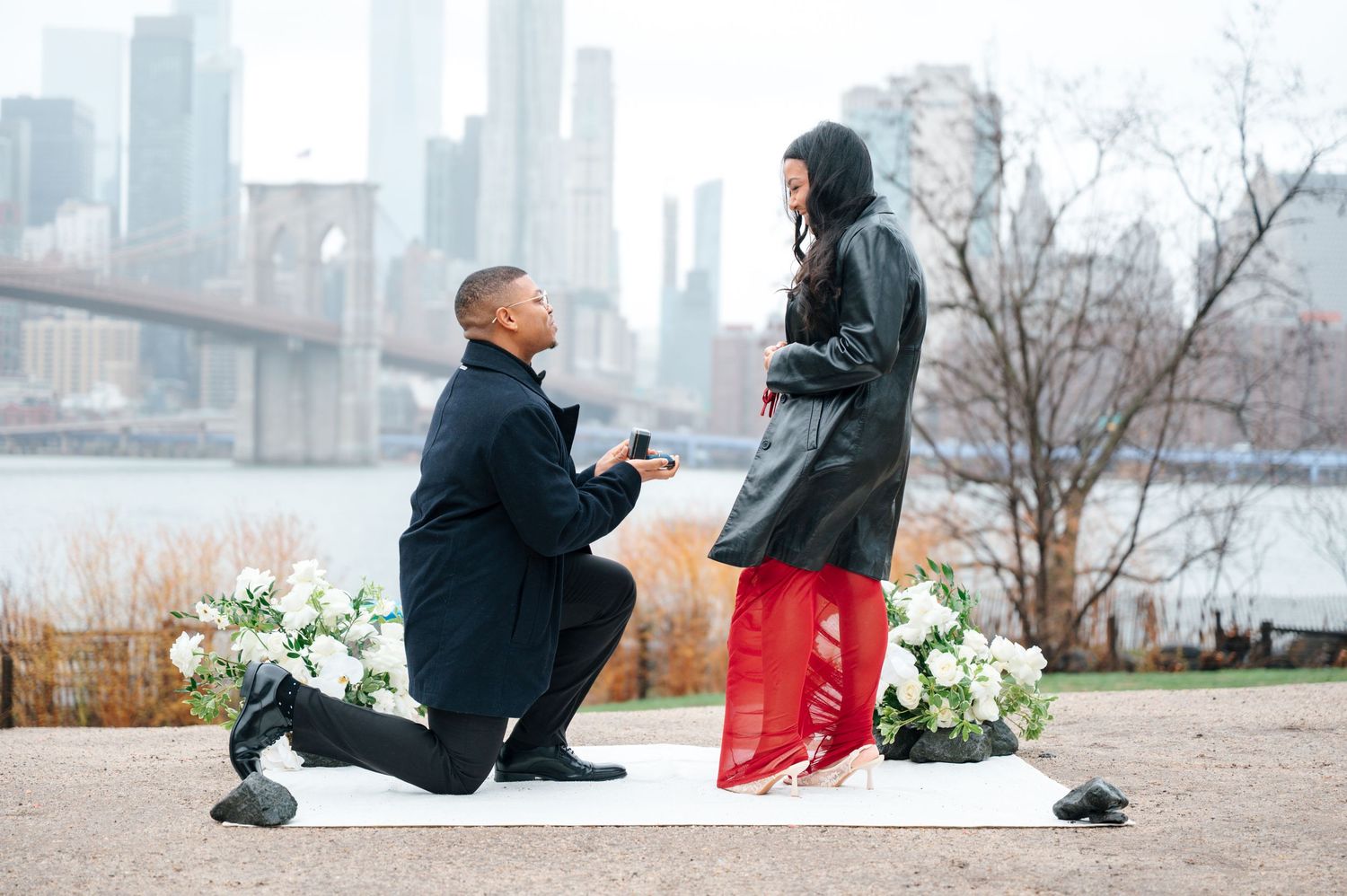 A romantic outdoor proposal scene with city backdrop, white floral arrangements and Brooklyn Bridge behind by NYC proposal photographer.