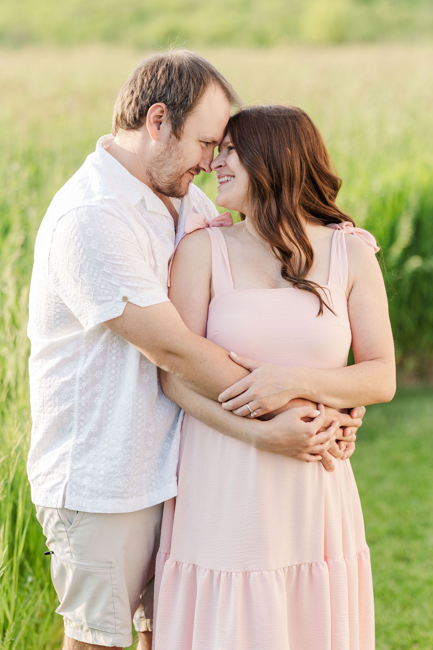 A couple shares an intimate moment together in a sunlit meadow wearing light colored clothes as they embrace.