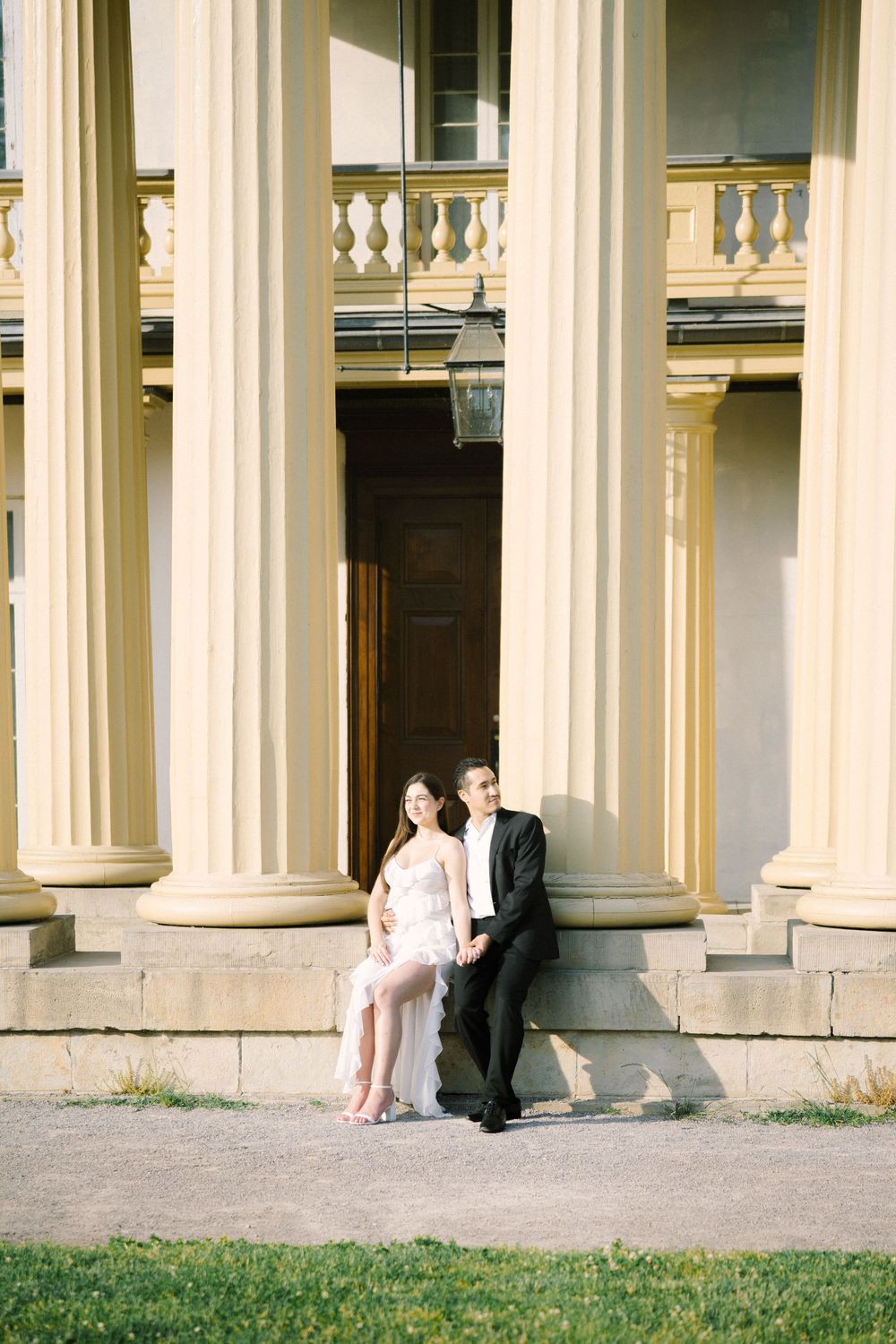 A couple sits between elegant white columns of a historic building during an artistic photoshoot.