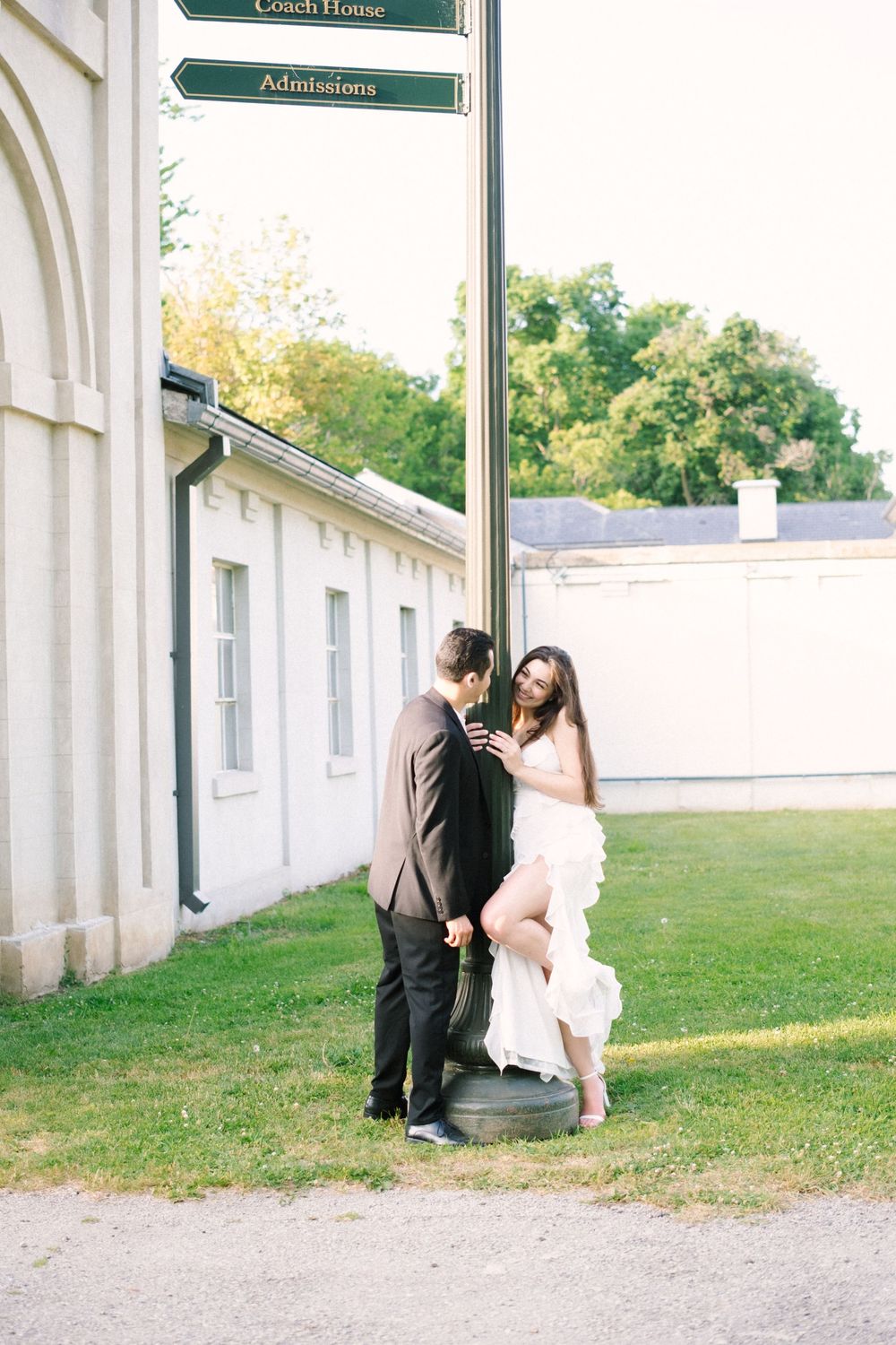 A couple in formal attire stands intimately on stone steps outside a white building with directional signs above.