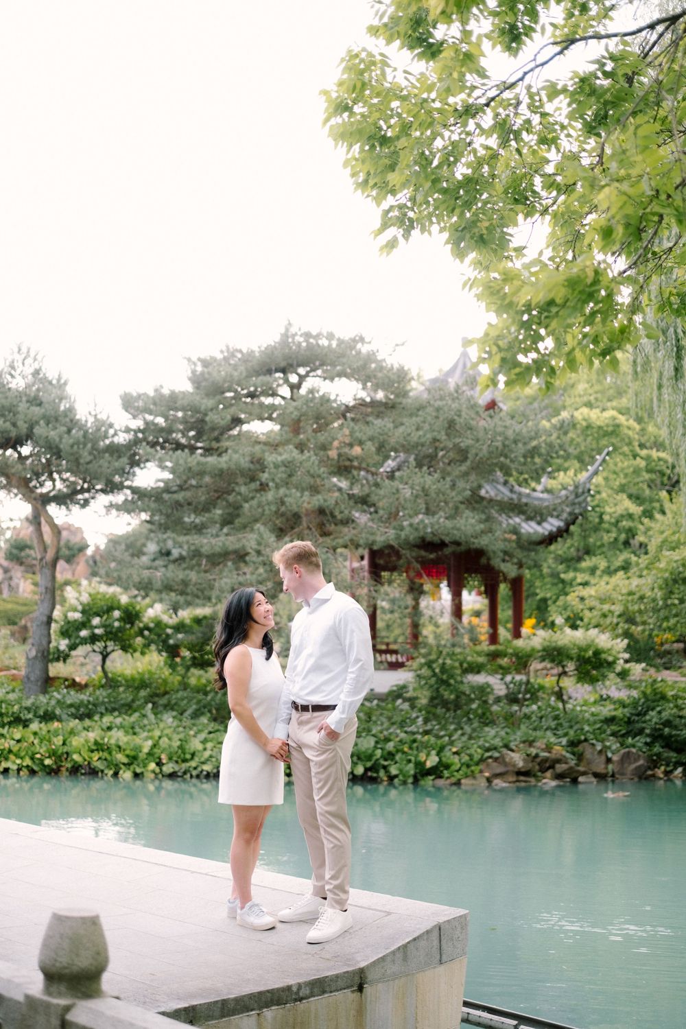 A couple standing together on a stone platform beside a tranquil turquoise pond in an Asian garden.