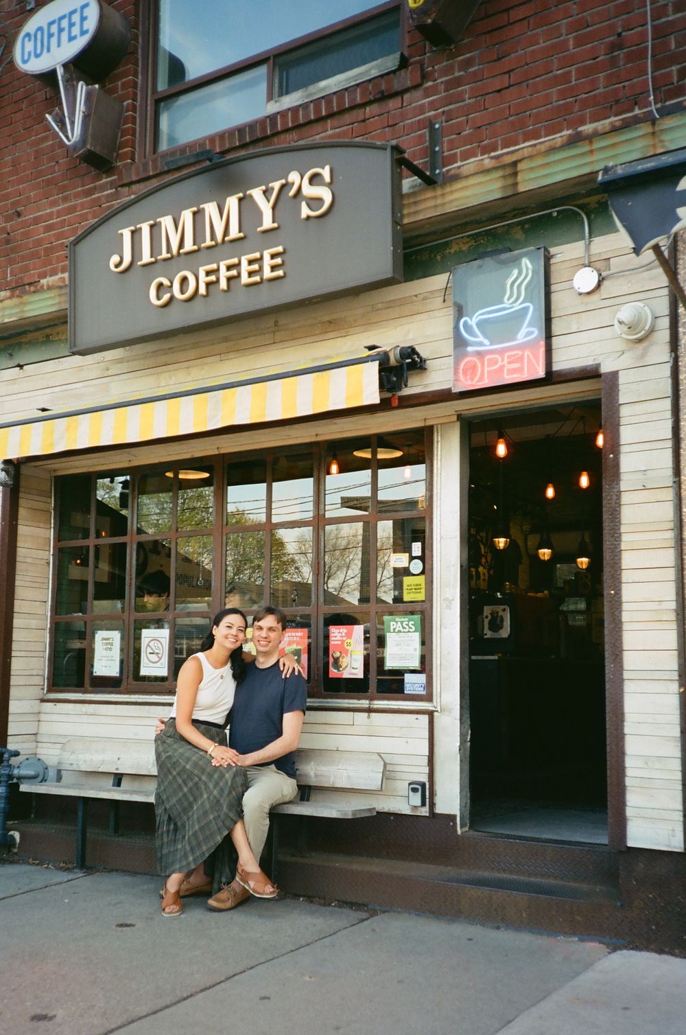 A couple sits together outside Jimmy's Coffee shop with a brick storefront backdrop.