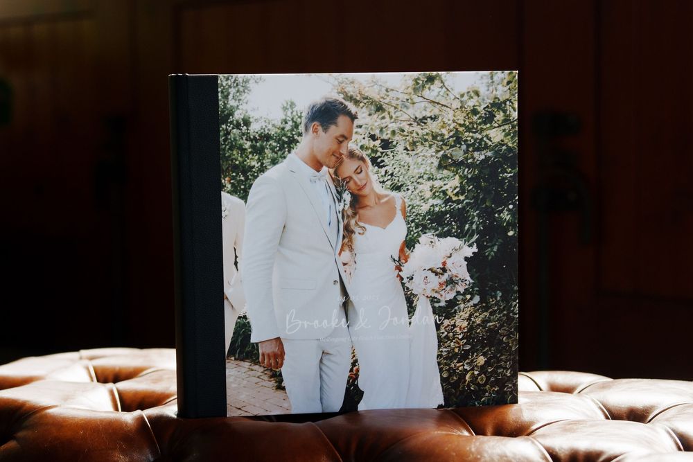 Hand holding a leather photo album displaying a romantic newlywed couple in white attire outdoors on their wedding day.