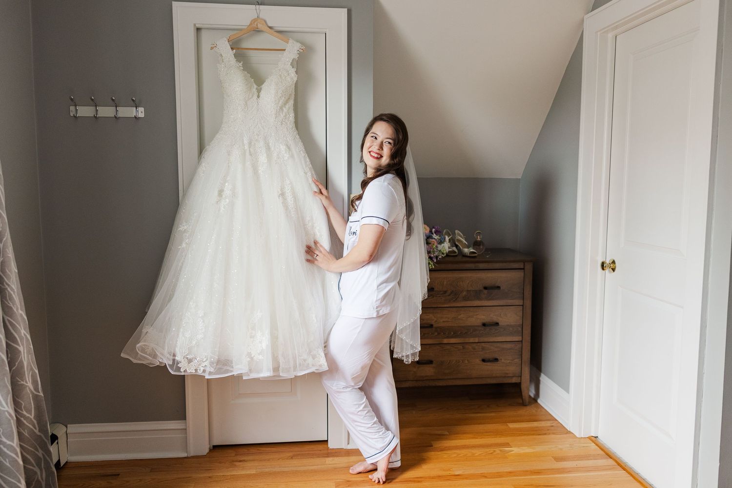 Person in white pajamas poses with an elegant white lace wedding gown hanging on display in a bedroom.