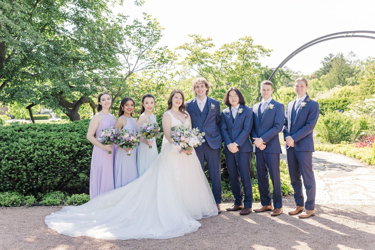 Wedding party in navy suits and lavender dresses poses together on a garden path on a sunny summer day.