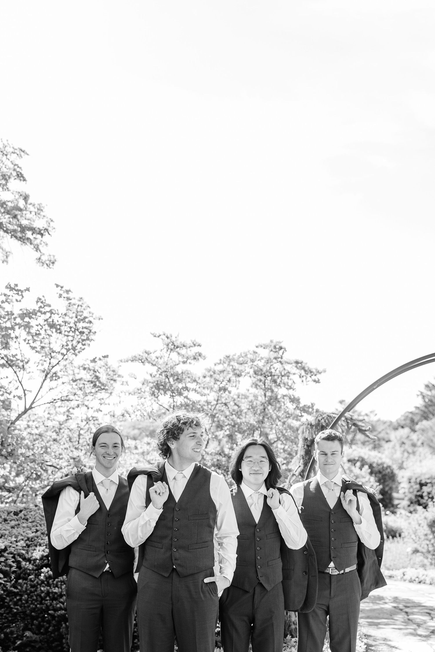 A black and white wedding party photograph showing four people in matching dark vests laughing together outdoors.