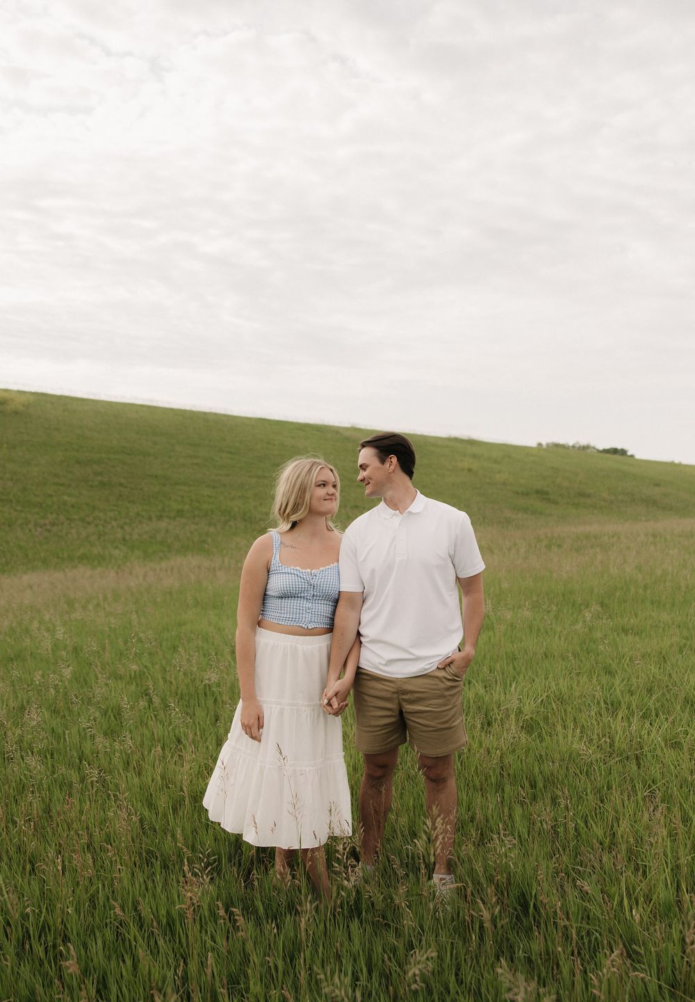 A romantic couple stands together in a green meadow under cloudy skies wearing casual summer attire.