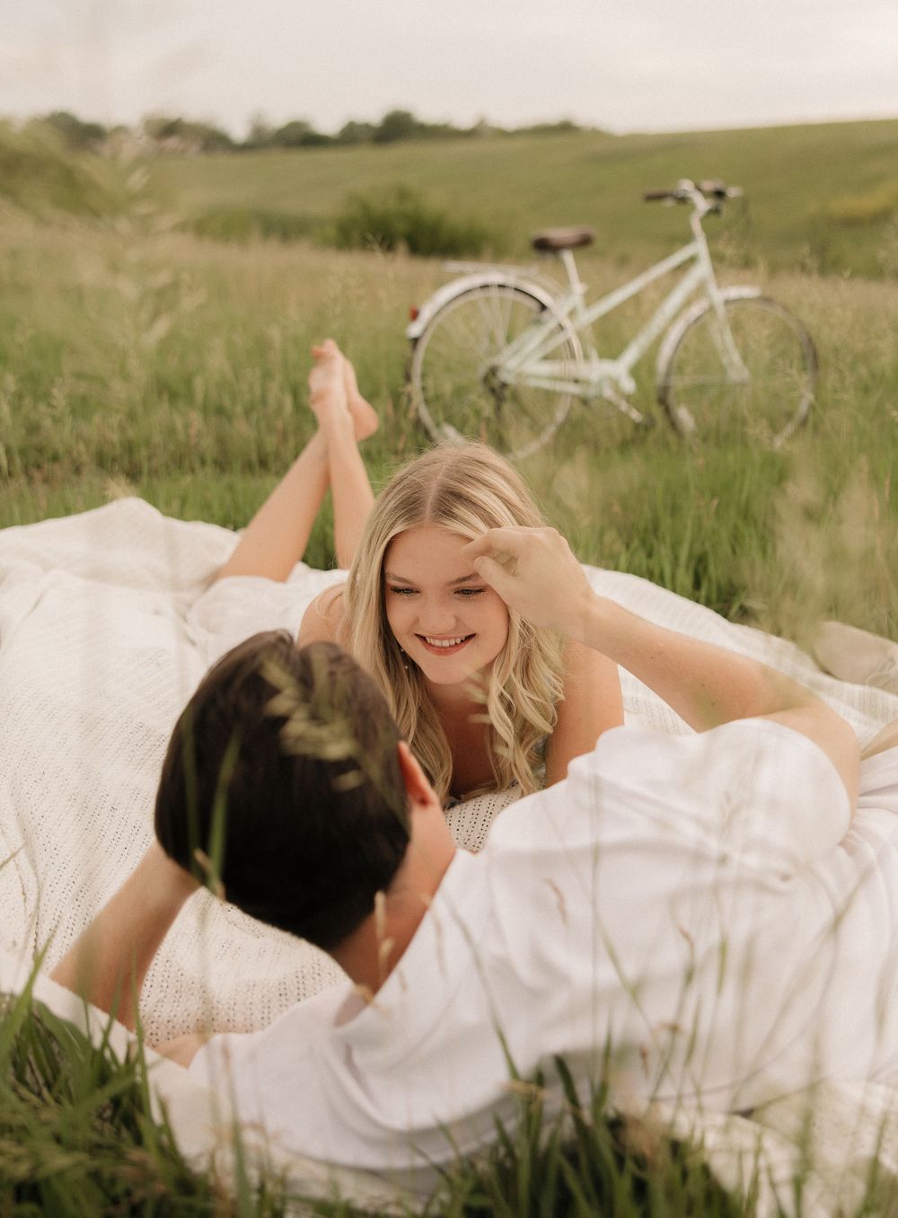 Two people share a romantic moment on a blanket in a grassy field with a white bicycle in the background.