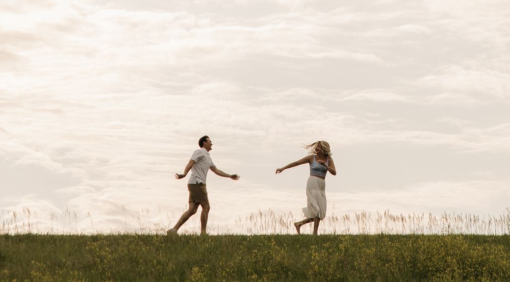 Two silhouetted people dance together in a grassy meadow at sunset against a cloudy sky.