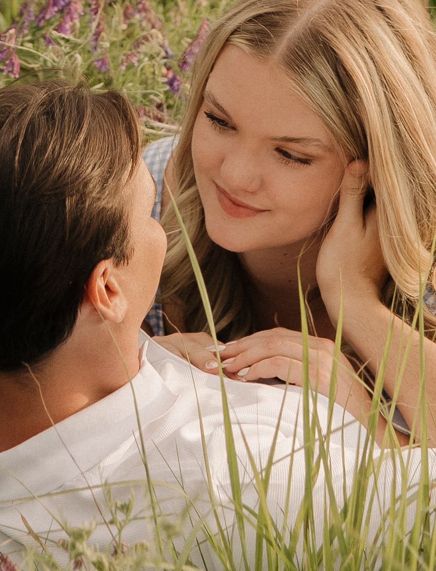 A romantic couple gazes at each other while lying in tall grass and wildflowers on a sunny day.