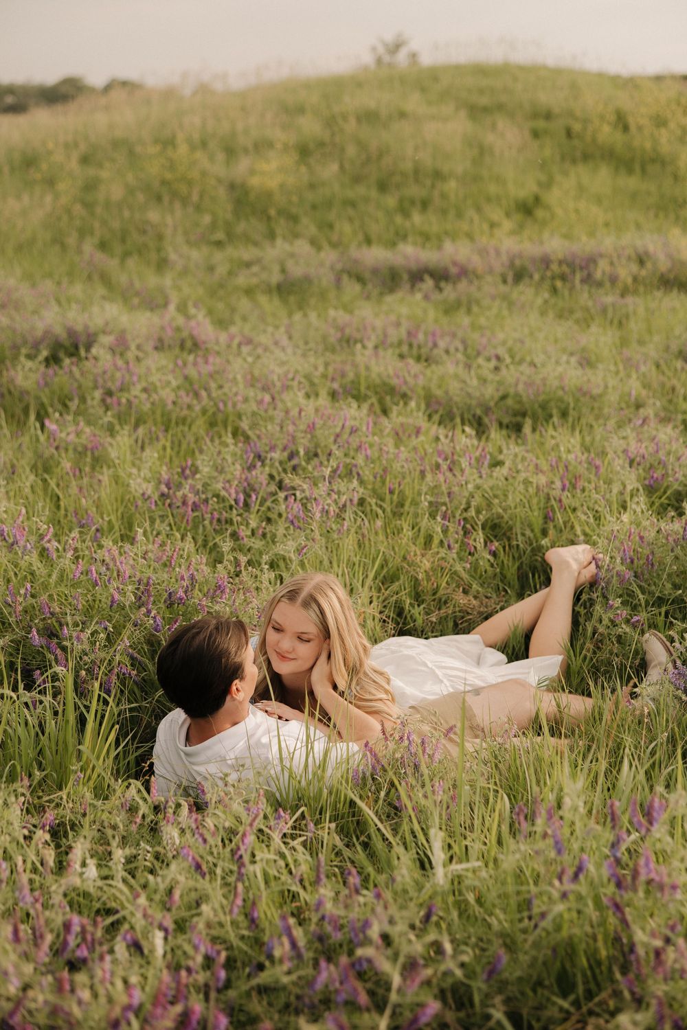 A couple laying together in a field of purple wildflowers on a sunny summer day.