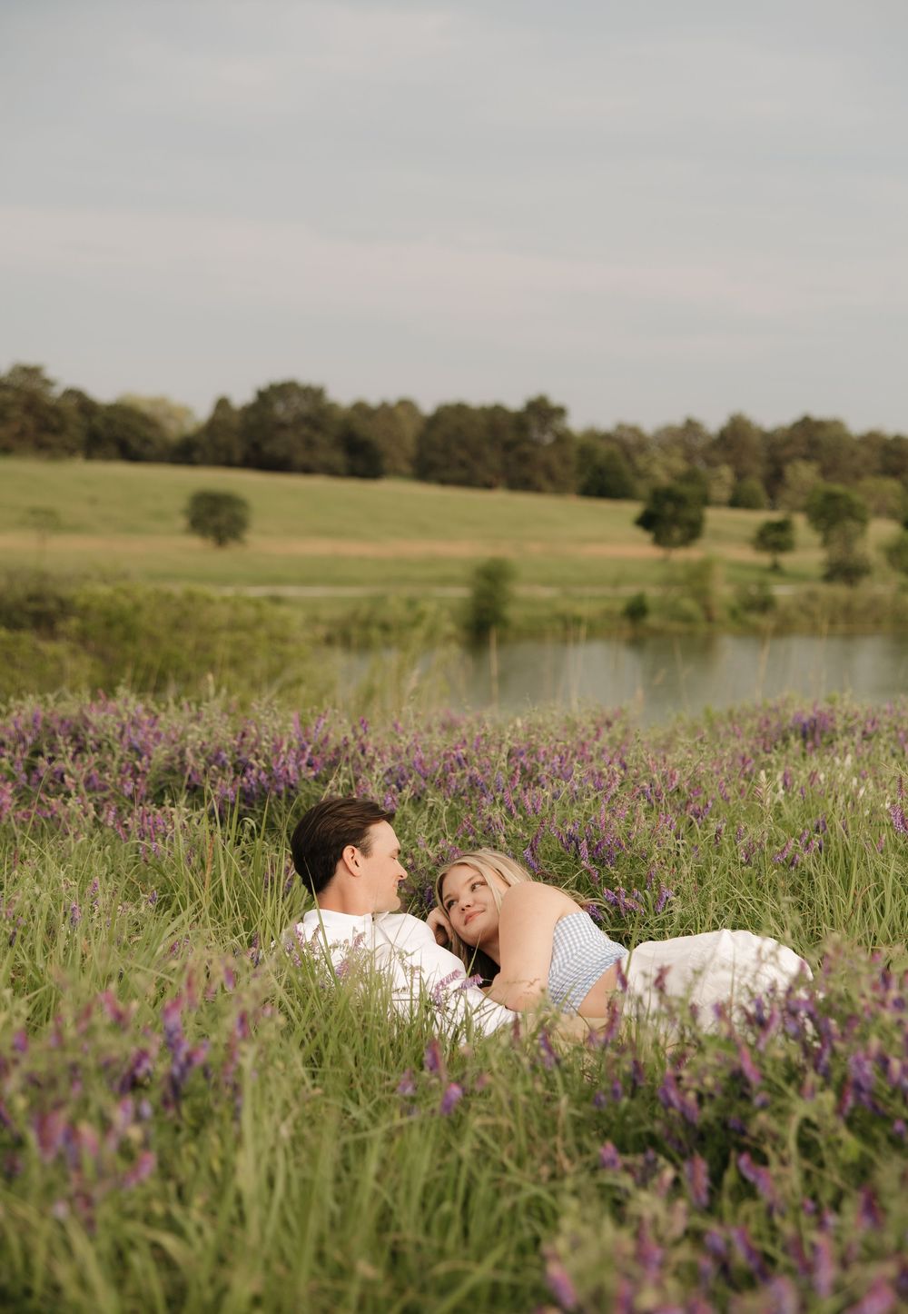 A romantic couple resting in tall grass and purple flowers near a pond.