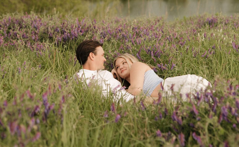 A couple rests together in a meadow filled with blooming purple flowers near a pond.
