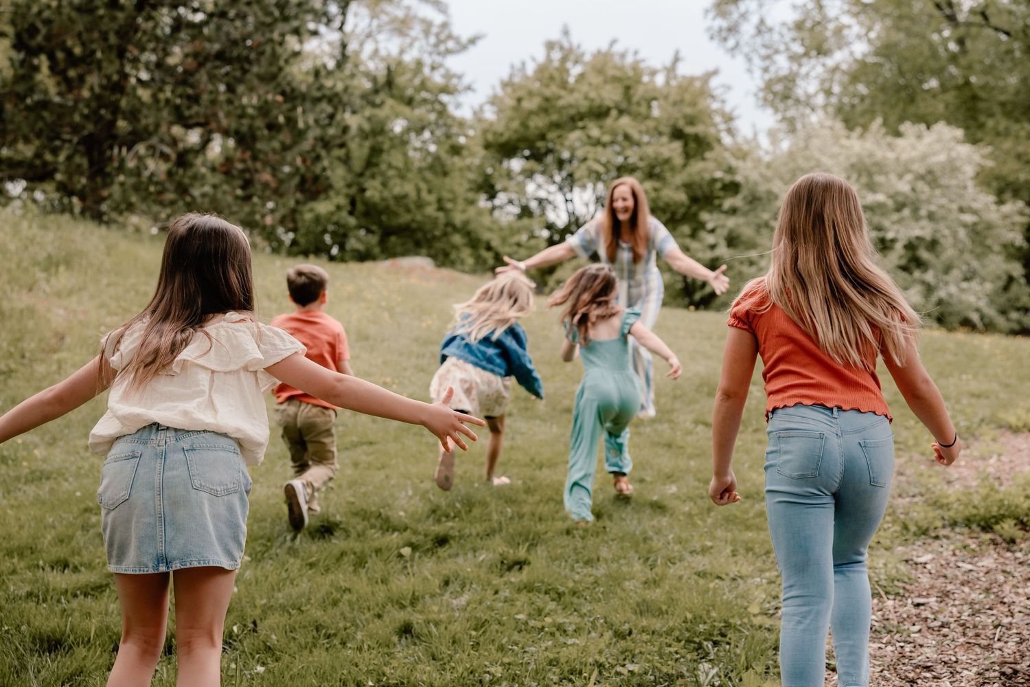 A group of people holding hands in a playful circle while dancing together on a grassy hill in summertime.