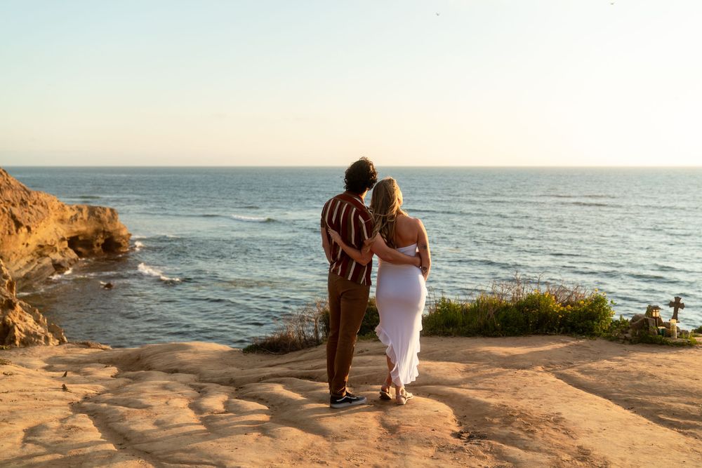 A couple stands together on a coastal cliff overlooking the ocean during golden hour.