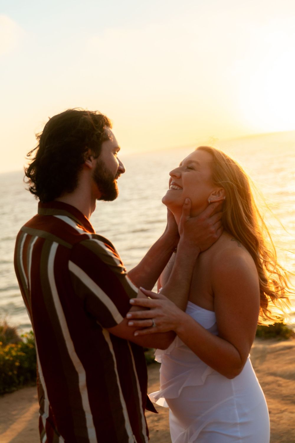 A couple embracing during sunset on a beach with a warm glow and ocean backdrop.