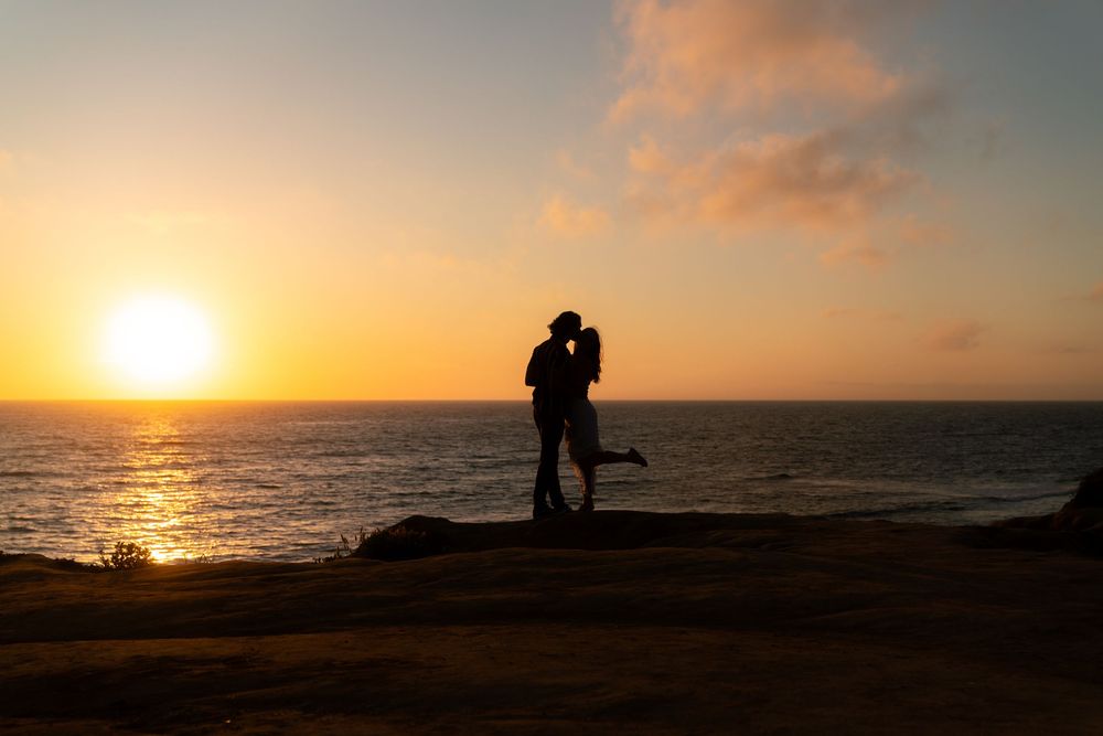 Silhouetted couple embracing on coastal cliff during romantic golden sunset over ocean horizon.