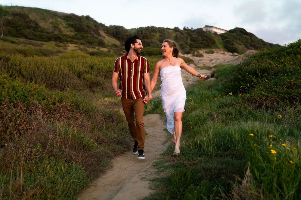 Two people in casual attire walk hand in hand along a scenic coastal trail surrounded by green vegetation at sunset.