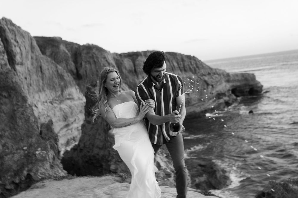 A couple shares a joyful moment on rocky cliffs by the ocean in black and white.