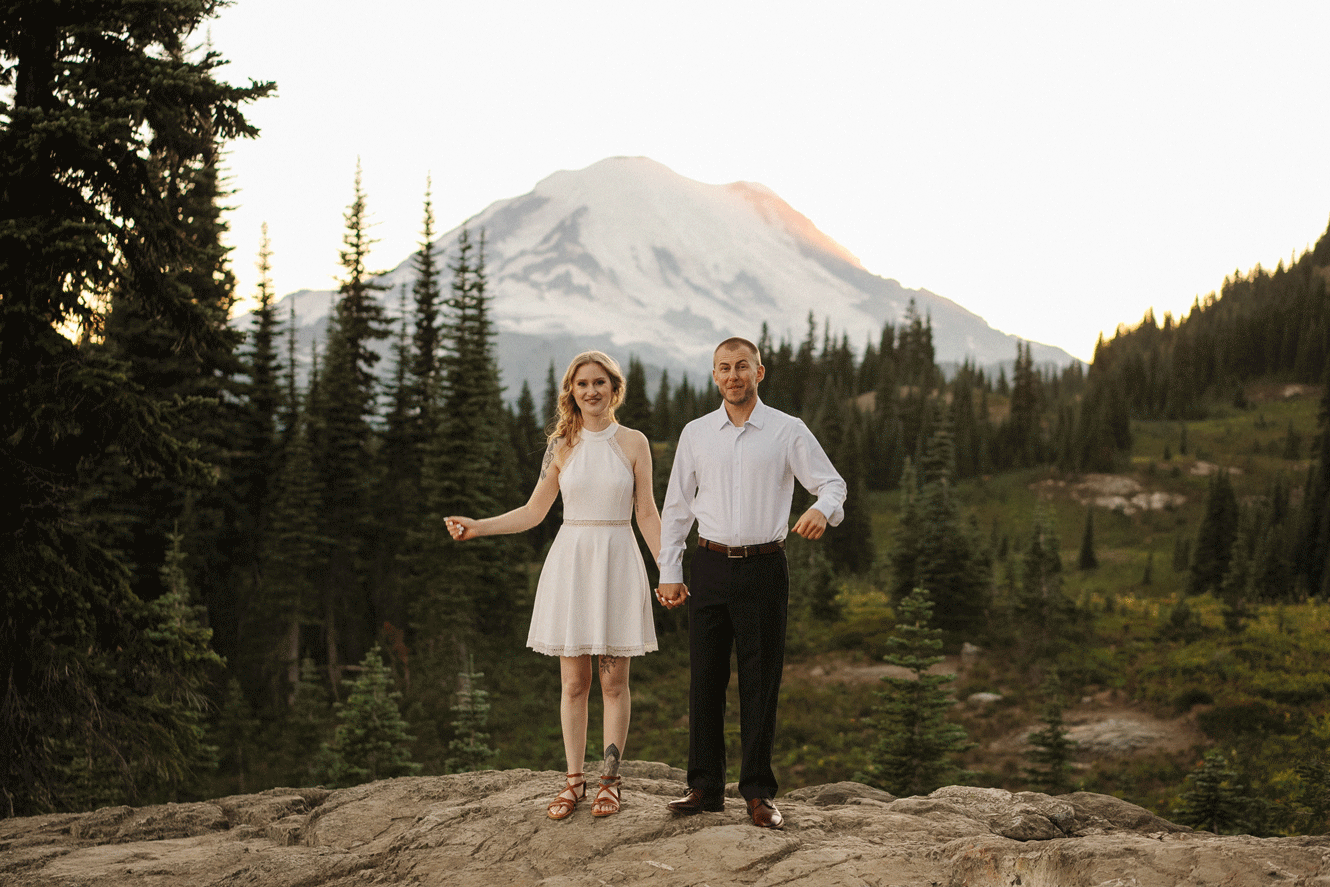 A couple stands on a rocky overlook with Mount Rainier's snow-capped peak rising majestically in the background.
