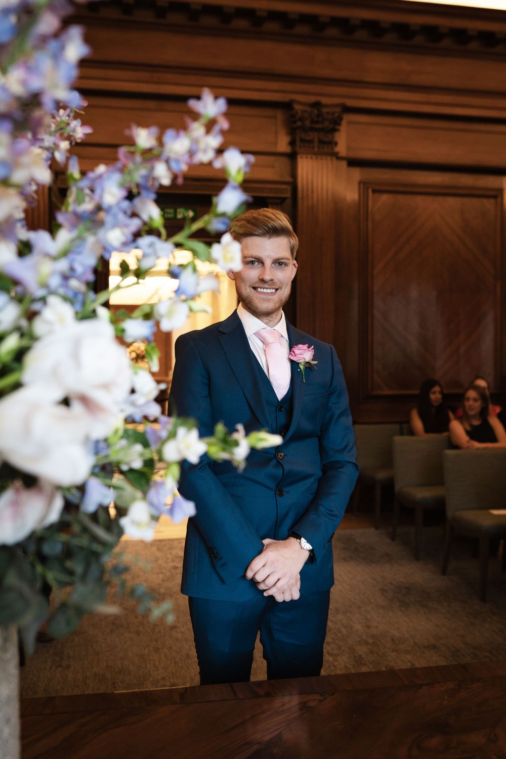 Wedding party members smile in elegant wood-paneled venue with floral decorations.